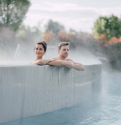 Paar in Kaskaden-Becken | © Spa Resort Geinberg / Chris Perkles Paar entspannt im Kaskaden-Becken, Dampf steigt auf und im Hintergrund sind farbige Herbstbäume | © Spa Resort Geinberg / Chris Perkles
