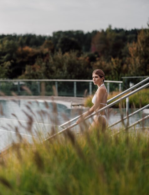 Blonde Frau im weißen Bikini betritt im Herbst das Kaskaden-Becken der Therme im Spa Resort Geinberg | © Spa Resort Geinberg / Chris Perkles