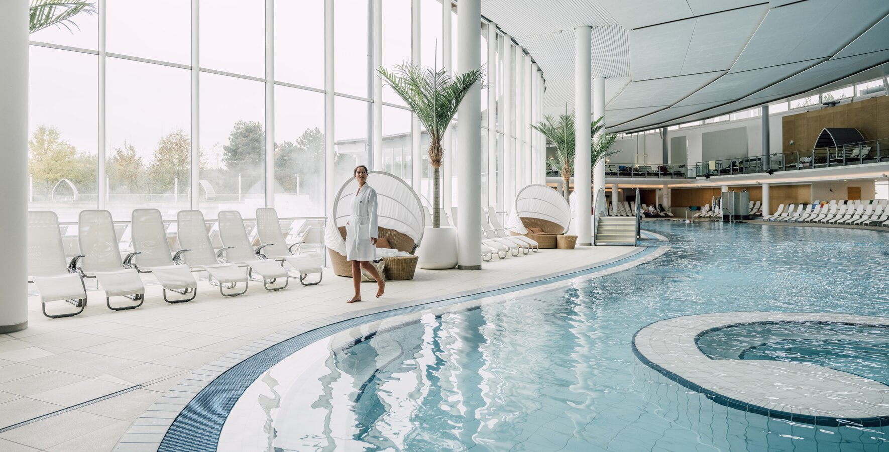 Großzügiger Innenbereich der Therme Geinberg | © Spa Resort Geinberg / Chris Perkles Frau im Bademantel schlendert am Rand des Thermal-Becken indoors in der weitläufigen Therme Geinberg | © Spa Resort Geinberg / Chris Perkles